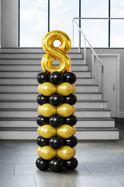 Black and gold stacked balloon column topped with a gold foil number balloon, displayed indoors near a staircase for a birthday or milestone celebration.